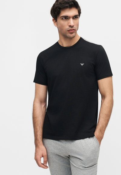 Άνδρας που στέκεται με τα χέρια στις τσέπες, φορώντας μαύρο κοντομάνικο μπλουζάκι με μικρό λογότυπο και ανοιχτό γκρι παντελόνι σε λευκό φόντο.