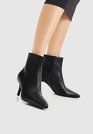 Bottines en cuir noir à bout pointu avec fermetures éclair latérales et talons hauts fins, portées avec des jambes nues et une jupe noire.