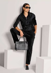 Vrouw in een zwart velour trainingspak, met zonnebril en loafers, houdt een zwarte leren handtas vast terwijl ze poseert met één voet op een witte kubus tegen een effen muur.