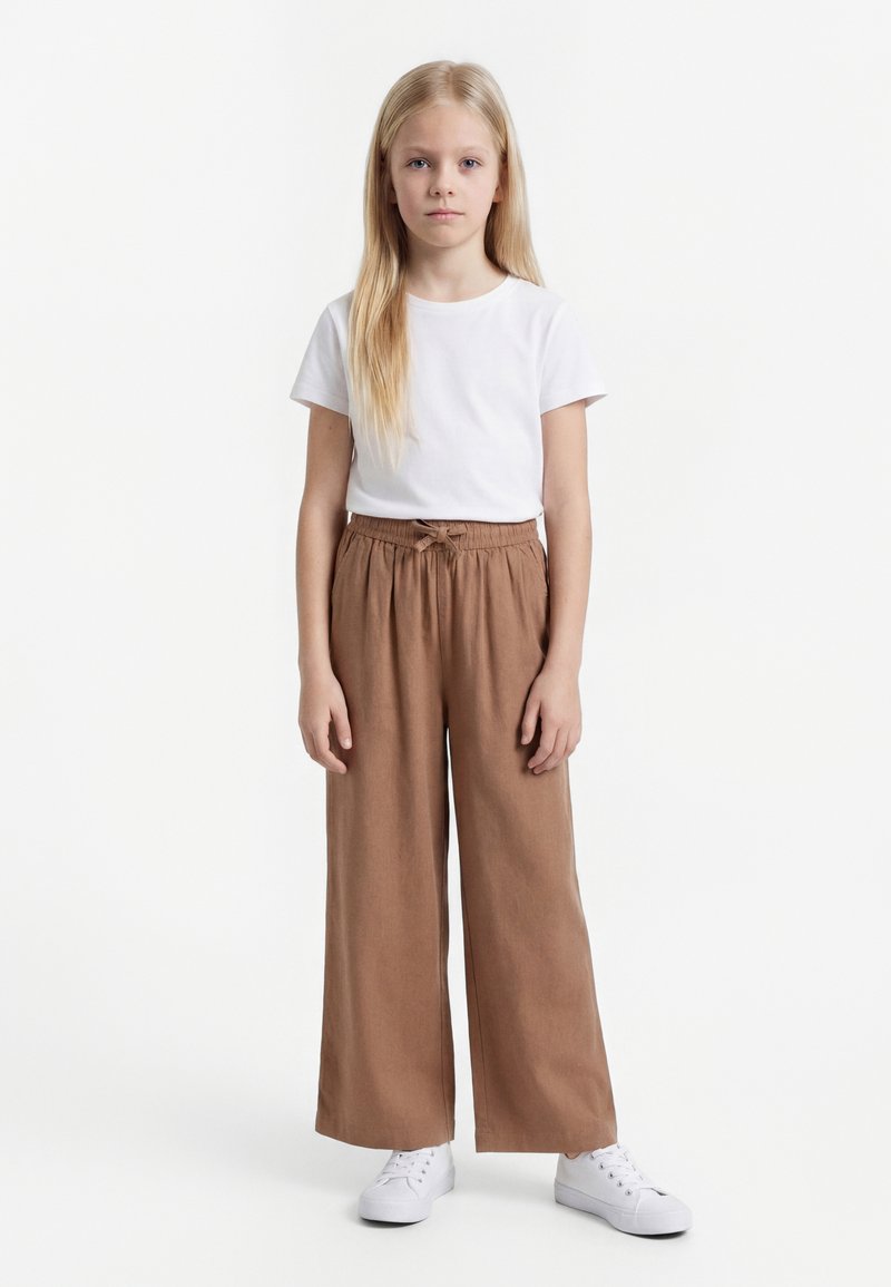 Ung pige med langt blondt hår iført en hvid t-shirt, brune vidde bukser og hvide sneakers, stående mod en ensfarvet hvid baggrund.