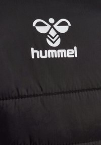 Svart pufferjacka med en mjuk textur och kviltade sektioner, med en framträdande vit "hummel"-logotyp och stiliserad bi-design.