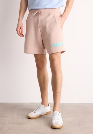 Homme portant un short beige avec un cordon turquoise et un texte bleu style graffiti sur la jambe gauche, associé à des baskets blanches sur un sol gris.