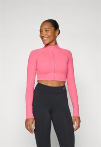 Fitted, cropped zip-up jacket in bright pink, made from stretchy material. Long sleeves and seamless design complement high-waisted black leggings.