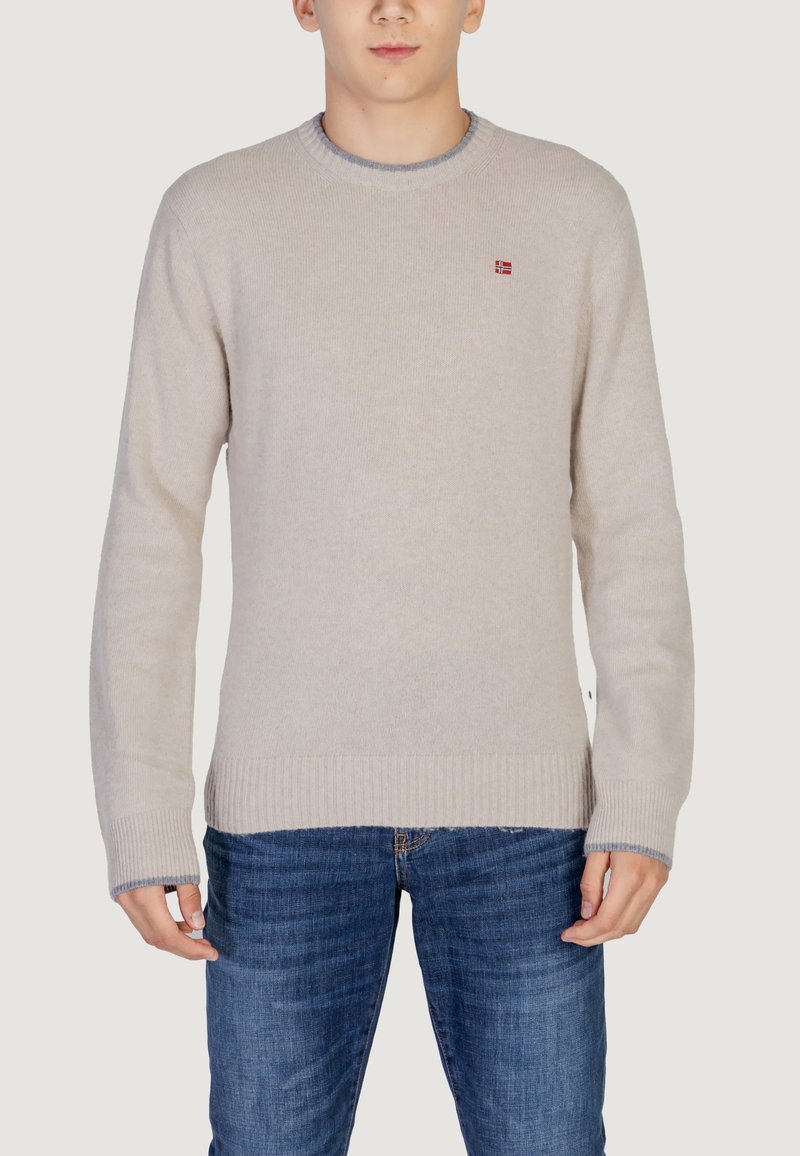 Beiger Pullover mit Rundhalsausschnitt und gerippten Ärmelbündchen. Verfügt über ein kleines rotes Logo auf der Brust. Getragen mit blauen Jeans. Weiche Textur.