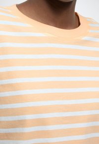 Tricou de culoare piersică pal cu dungi orizontale albe, cu guler rotund și țesătură moale și texturată. Vedere apropiată a gulerului și modelului.