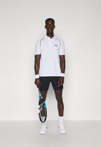 Polo blanc avec des accents jaunes, shorts noirs et chaussures de sport blanches. Le mannequin tient une raquette de tennis, mettant en avant une coupe athlétique et un design sportif.