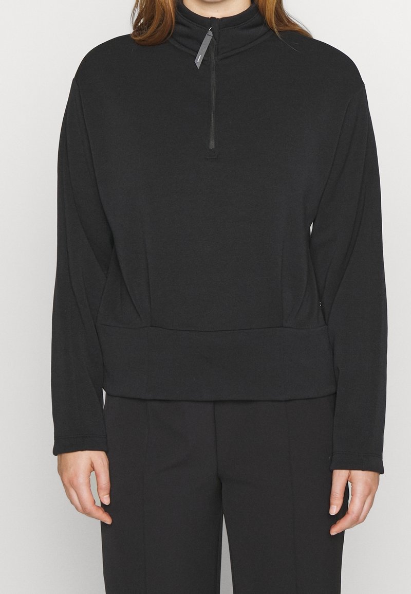 Personne portant un pull noir à manches longues avec une fermeture éclair quart de zip et un col haut, associé à un pantalon noir, debout devant un fond uni.