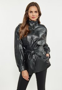 Giacca in pelle nera con colletto alto, chiusura con bottoni, due tasche sul petto, vita con cintura e maniche lunghe. Texture liscia e vestibilità su misura.