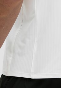 Biela atletická tričko so štruktúrovaným povrchom, krátkymi rukávmi a detailmi na bočných švoch. V spodnej časti doplnené čiernymi šortkami.