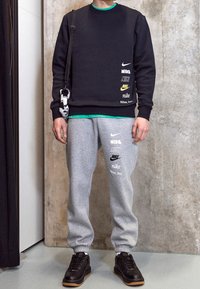 Svart sweatshirt med logotryck, grå sweatpants med varumärke, och svarta sneakers med tan-färgade sulor. Modellen poserar mot en betongvägg.