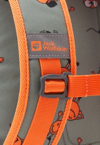 Détail d'un sac à dos Jack Wolfskin avec un tissu vert présentant des graphismes de dessins animés orange, des bretelles orange vif et une étiquette grise avec le logo.