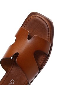 Sandalo in pelle marrone con un design traforato. Presenta una suola piatta, un bordo cucito e una superficie liscia con una larga fascia che attraversa il piede.