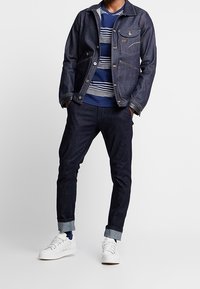 Jeansjacka med kontrasterande stygn, randig blå och vit tröja med rund hals, mörka åtsittande jeans och vita sneakers med uppvikta ben.