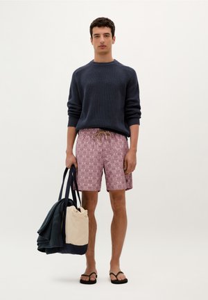 Pulover tricotat navy, pantaloni scurți modelați roz cu șnur, șlapi negri, ținând o geantă tote navy și bej. Ținută simplă și casual.