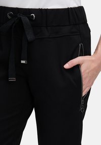 Zbliżenie czarnych spodni z elastycznym pasem ze sznurkiem i boczną kieszenią na zamek, z ręką częściowo wsuniętą w kieszeń.