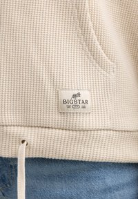 Hanorac de culoare crem, dintr-un material texturat, cu model waffle-knit. Dispune de o buzunar frontal și un eticheta cu "BIGSTAR" pe tivul inferior.