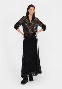 Donna che posa con giacca nera trasparente in pizzo floreale con cerniera e gonna lunga coordinata in pizzo, indossando scarpe da ginnastica nere su sfondo bianco.