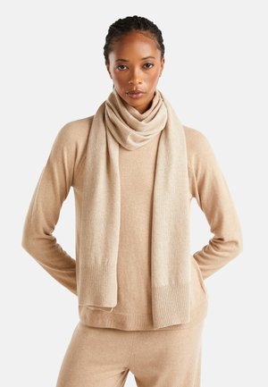 Pull en cachemire beige avec des poignets côtelés, associé à une écharpe assortie drapée autour du cou. Texture douce et coupe ajustée.