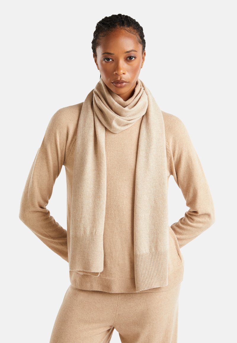 Maglione beige in cashmere con polsini a coste, abbinato a una sciarpa coordinata appoggiata intorno al collo. Texture morbida e vestibilità su misura.