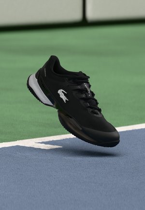 Baskets noires Lacoste avec logo crocodile blanc, flottant au-dessus d'une surface de terrain de tennis bleu et vert près de la ligne blanche de délimitation.