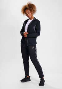 Zwarte sportjack met grijze accenten, gecombineerd met zwarte joggers met een chevronpatroon. Model draagt zwarte sneakers en een wit shirt.