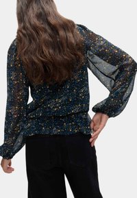 Luftigt sort blomstret bluse med lange, gennemsigtige ærmer, samlet talje og en mørkegrøn bund. Har små blå og guld blomstermønstre.