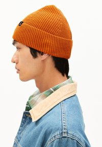 Orange ribbed beanie with a peak at the top, worn by a person in profile. Background is plain white, showcasing the hat’s texture and color.