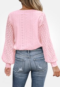 Mujer con una blusa de encaje rosa de mangas largas abullonadas y jeans de mezclilla azul desgastados, vista desde atrás sobre un fondo blanco.