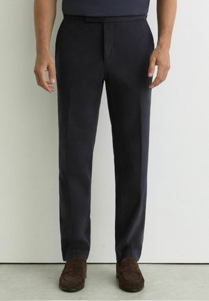 Mężczyzna w ciemnogranatowych, dopasowanych spodniach i brązowych zamszowych mokasynach, stojący na tle jednolitej jasnej ściany i podłogi.