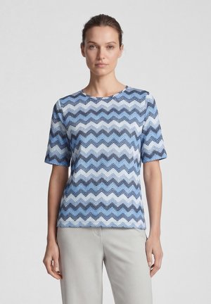 Woman wearing a blue and white zigzag patterned short-sleeve top with light gray pants, standing against a plain background.