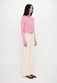 Gilet court rose avec fermeture à boutons et deux poches poitrine, associé à un pantalon large blanc et des chaussures plates marron.
