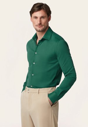 Man draagt een groen overhemd met lange mouwen en beige broek, met handen in de zakken, poserend tegen een effen lichte achtergrond.