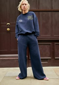 Navy blue sweatshirt dengan cetakan "New York" dan nomor "85," dipadukan dengan celana lebar navy dan sepatu slip-on pink. Tekstur kain yang lembut.