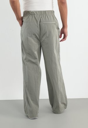Personne portant un pantalon ample gris clair avec une ceinture élastique, debout sur un sol gris devant un fond blanc, vue de dos.