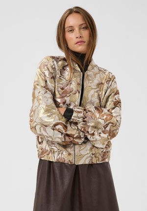 Joven mujer con cabello largo y castaño lleva una chaqueta bomber metálica con flores sobre una camiseta negra y una falda marrón oscura, de pie con los brazos cruzados.