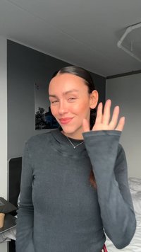 Šedý rebrovaný top s dlhým rukávom a kapucňou, s širokými manžetami. Osoba sa usmieva a máva, na neutrálnom pozadí a v jemnom osvetlení.