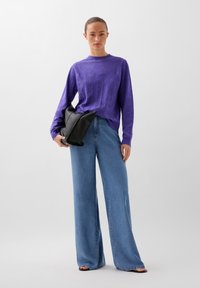 Lilla strikket sweater med et struktureret mønster, parret med lyseblå brede jeans. Modellen holder en sort læder clutch.