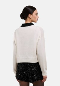 Pull court crème avec un col noir, présentant une texture douce et une coupe décontractée. Associé à une jupe noire à paillettes.