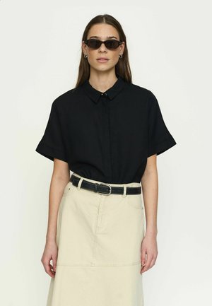 Kobieta w czarnej koszuli z krótkim rękawem i kołnierzykiem, beżowej spódnicy z wysokim stanem z czarnym paskiem oraz kanciastych czarnych okularach przeciwsłonecznych, stojąca na tle białego tła.