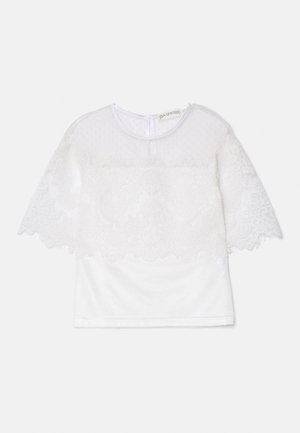 White top featuring a textured sheer lace overlay with scalloped edges and floral patterns. Short sleeves and a round neckline.