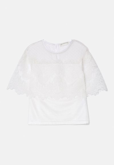 Hvid top med et tekstureret, gennemsigtigt spidsdæksel med bølgede kanter og blomster mønstre. Korte ærmer og rund halsudskæring.