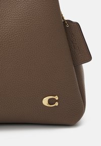 Barna texturált bőr kézitáska sarka arany "C" logóval és hozzákapcsolt bőr címtáblával, amely egy kis arany láncon lóg.