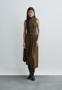 Brown ribbed sleeveless dress with a high neck, featuring pleated skirt, asymmetrical draping, and black knee-high boots as accessories.