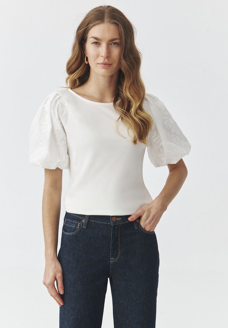 Top bianco con maniche corte e arricciate ricamate, abbinato a jeans in denim scuro. Design semplice e aderente con scollatura rotonda.