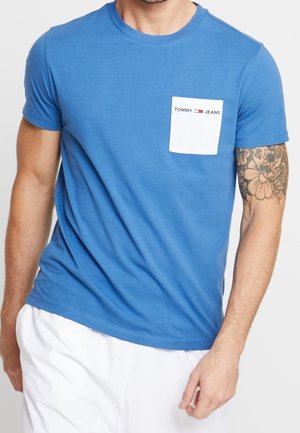 Man wearing a blue short-sleeve t-shirt with a white chest pocket labeled "Tommy Jeans" and white pants.