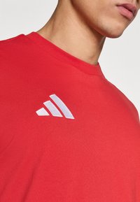 Κόκκινο βαμβακερό μπλο υζάκι με στρογγυλή λαιμόκοψη, κοντά μανίκια και λευκό κεντημένο λογότυπο με τρεις ριγέ λεπτομέρειες στο αριστερό στήθος.