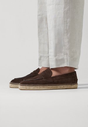 Mocassins en daim marron à enfiler avec semelles tissées style espadrille, portés avec un pantalon en lin clair.