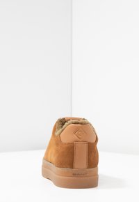 Baskets en daim marron avec une doublure en fausse fourrure, présentant un patch de talon en cuir beige et une semelle en caoutchouc transparent. Détail du logo doré à l'arrière.