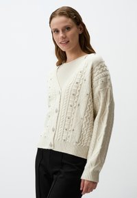 Crème kabelgebreide cardigan met V-hals, voorzien van textuurpatronen en versieringen, evenals een ribgebreide onderrand en manchetten.
