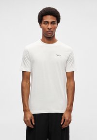 Tricou alb cu mâneci scurte din bumbac, cu guler rotund. Prezintă un mic logo negru pe partea stângă a pieptului. Textură netedă; croială lejeră.